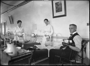 Two woman hospital workers stand behind bandaged children in beds, and a man sits next to them