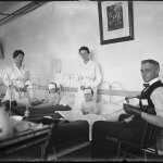 Two woman hospital workers stand behind bandaged children in beds, and a man sits next to them